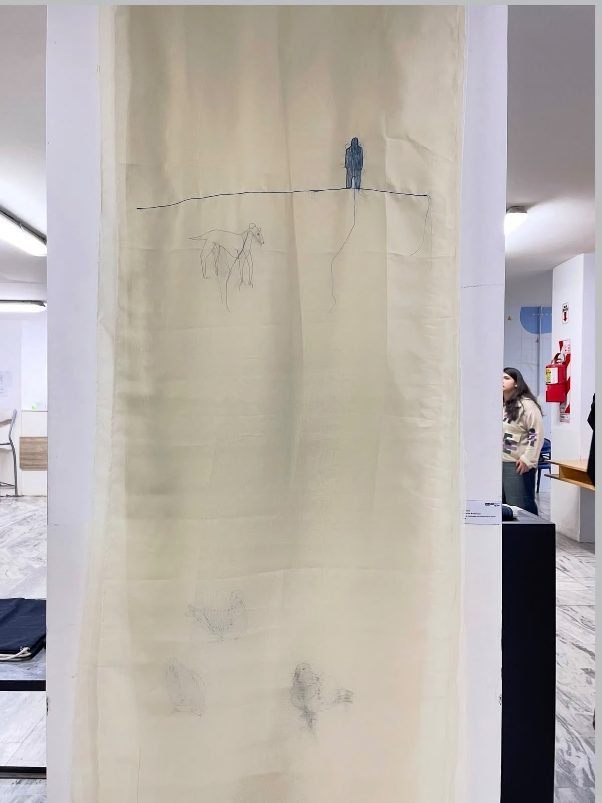 Obra textil colgada en la sala de la EMAC: tela traslúcida con una figura bordada en azul sobre una línea de horizonte